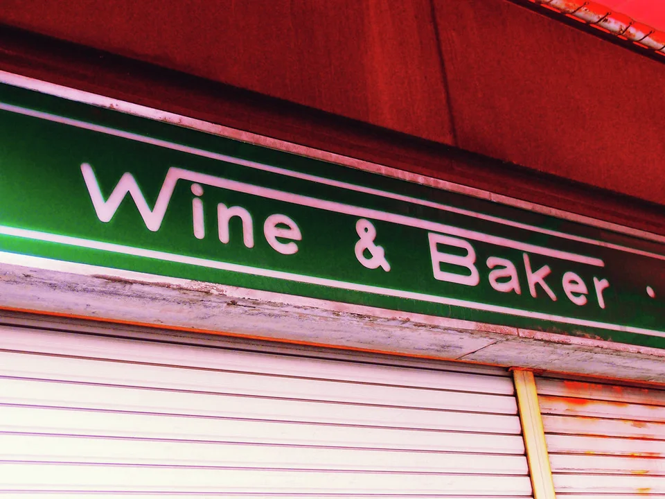 Pale wine and baker lettering runs across a dark green sign above closed white shutters.