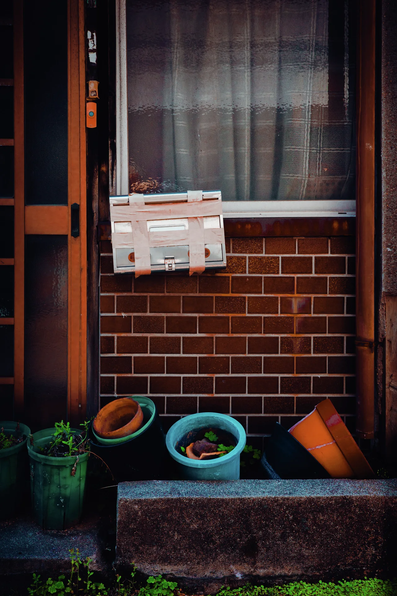 A pale mailbox hangs below a curtained window above colorful pots on a brick-fronted wall.