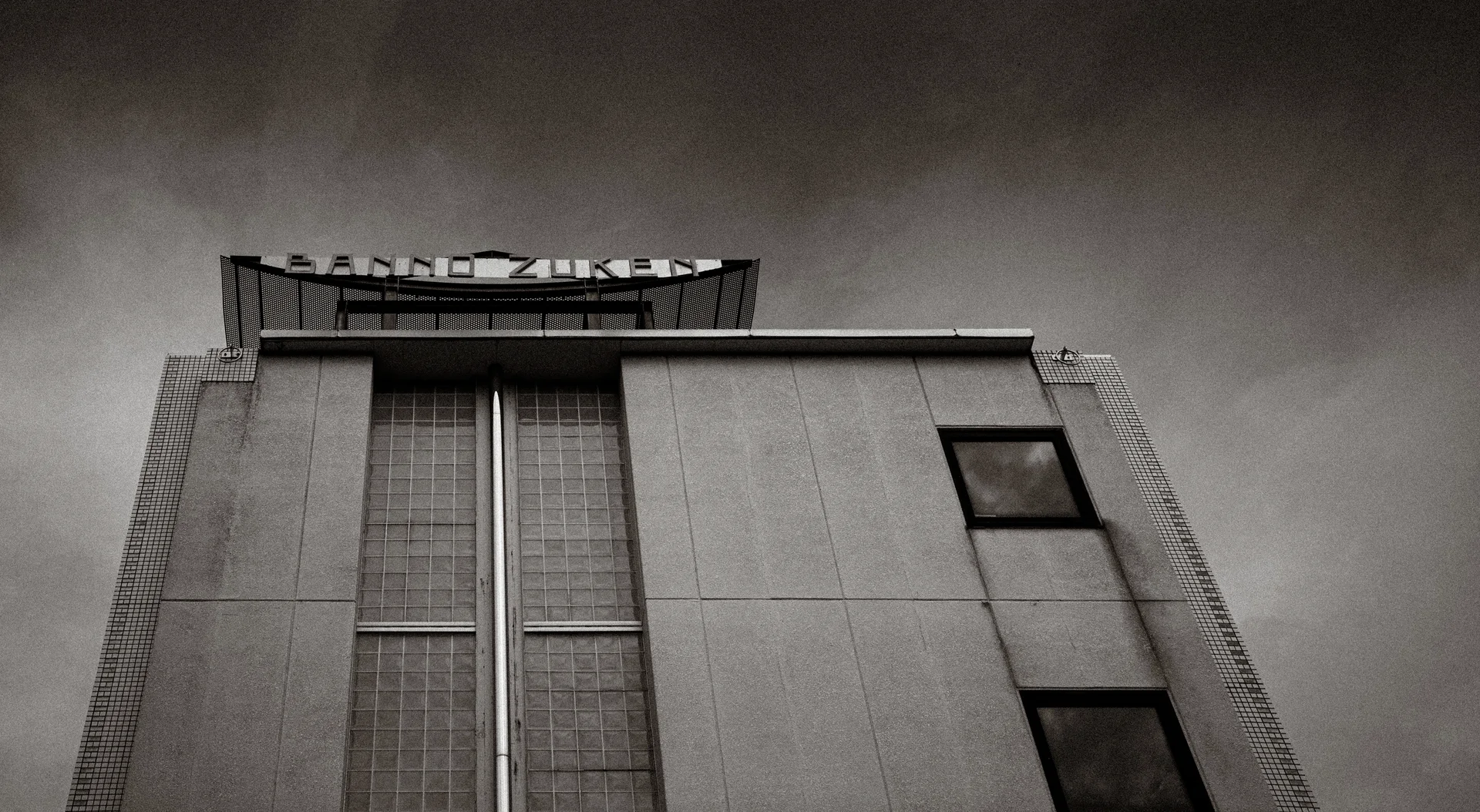A plain building rises beneath dark clouds, with narrow vertical windows and rooftop framework in black and white.