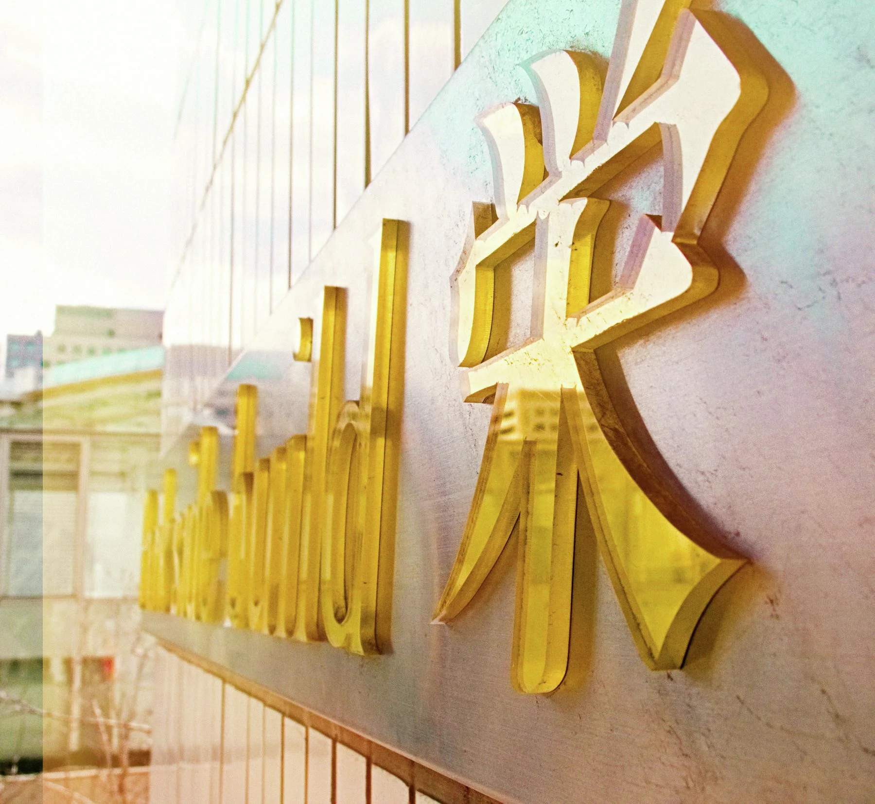 Thick gold lettering projects from a pale wall and casts sharp shadows in angled sunlight.