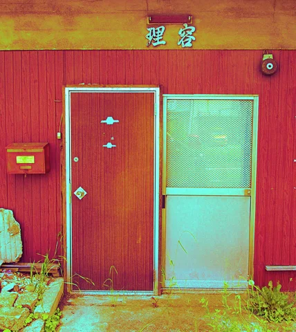 A plain reddish facade has two narrow doors, a small mailbox, and faded Japanese lettering above.