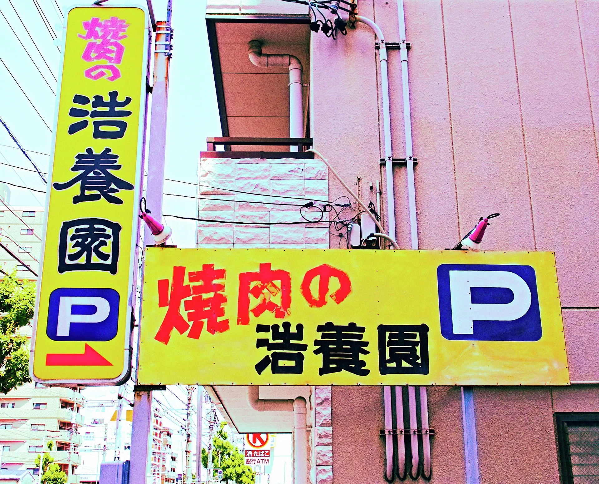 Bright yellow restaurant signs with red and black Japanese lettering display blue parking symbols beside a pink building.