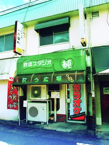 Green karaoke shop awnings and signs surround a small entrance partly blocked by an outdoor air conditioner.