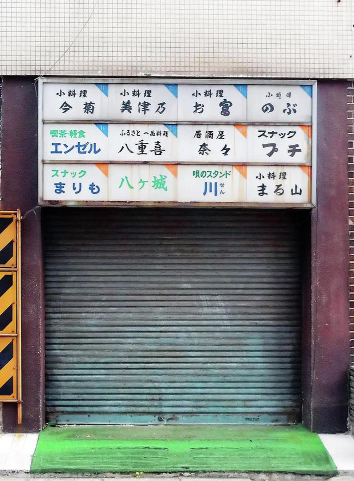 A grid of small business-name panels hangs above a closed dark shutter and green entrance mat.