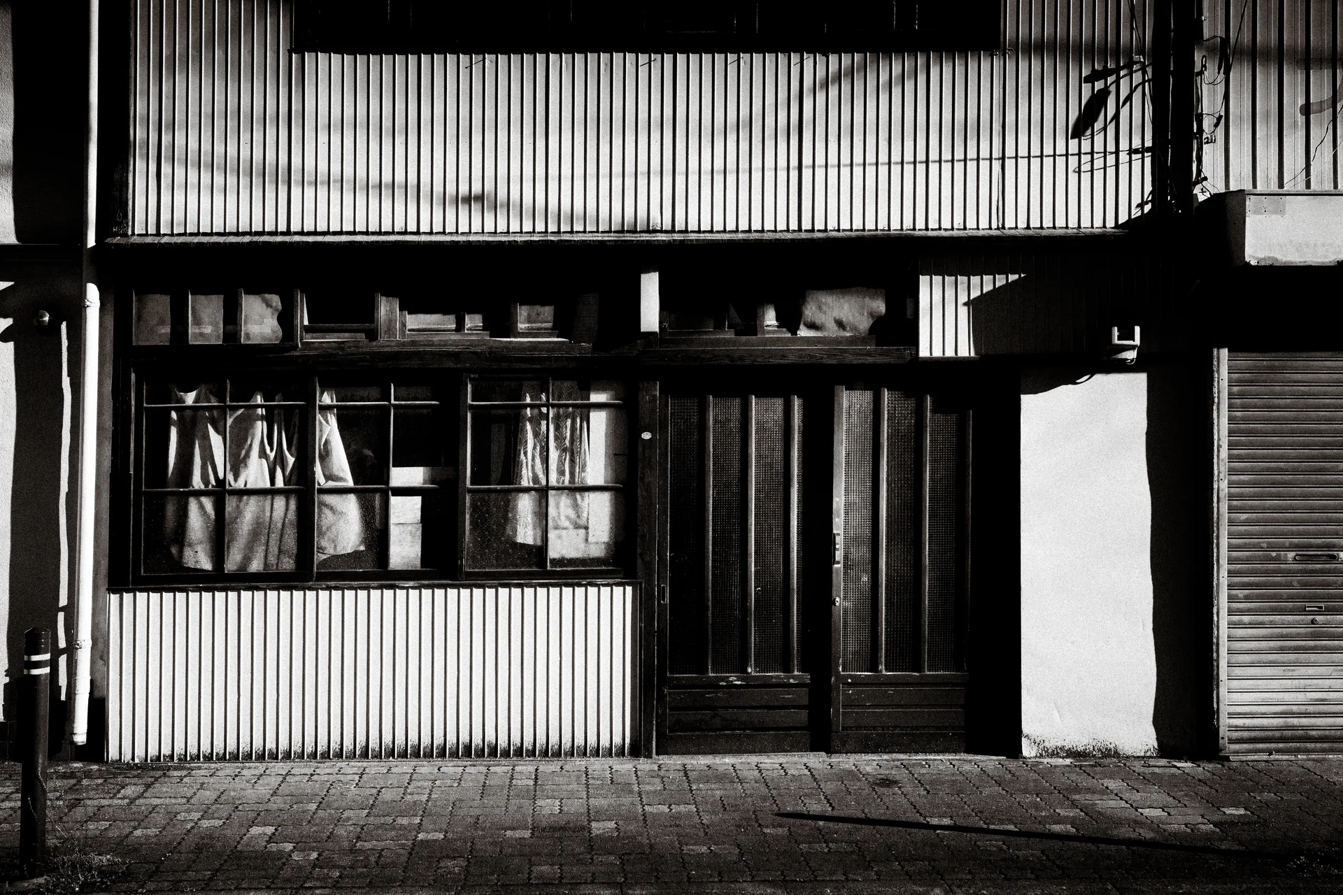 Curtained windows and wooden double doors interrupt a corrugated storefront above brick paving in black and white.
