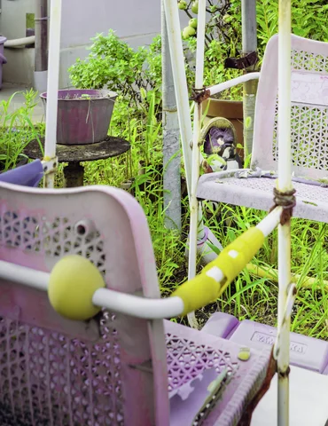 Perforated pale chairs and a yellow handlebar sit among plants and rusted supports.