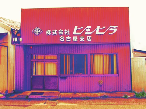 White Japanese lettering spans a red corrugated building above a narrow door and curtained windows.