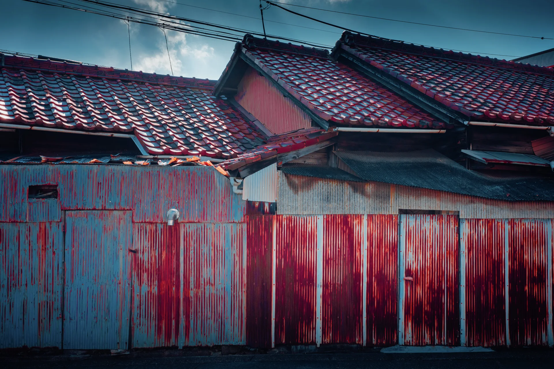 Red rust streaks cover blue and pale corrugated panels beneath layered dark tiled roofs.
