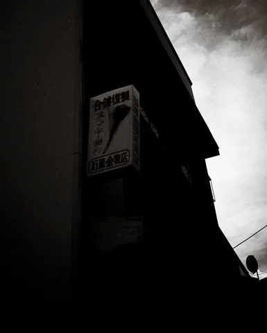 A projecting sign hangs from a deeply shadowed building beneath a narrow strip of cloudy sky.