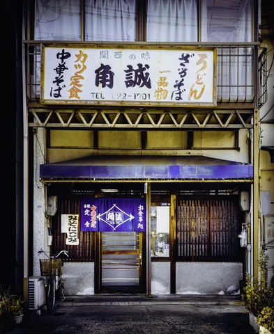 A narrow restaurant facade has a large Japanese sign, purple entrance curtains, and glass sliding doors.
