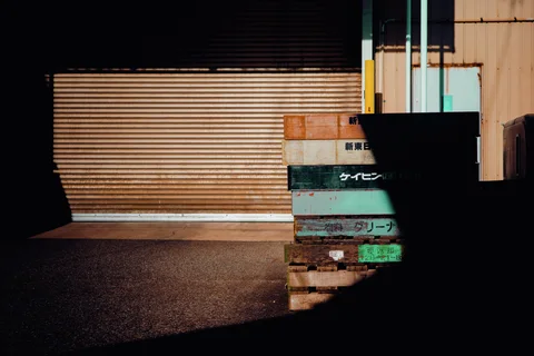 Sunlight forms a broad bright patch across a closed shutter beside stacked weathered wooden pallets.