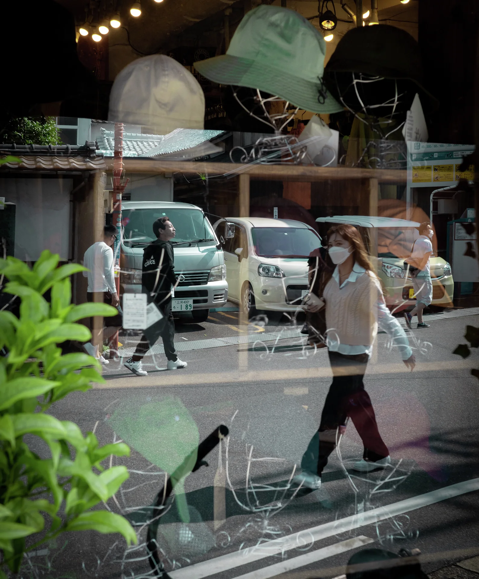 Pedestrians and parked vehicles reflect across a shop window displaying pale hats above leafy plants.