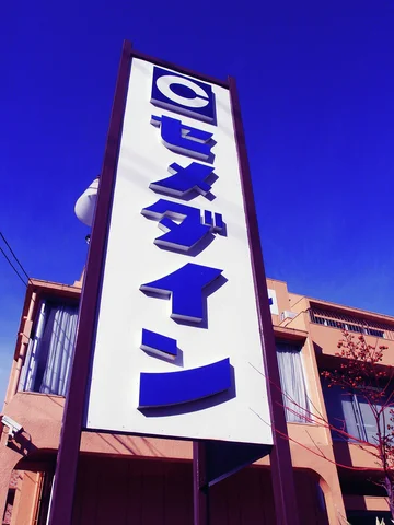 Large blue Japanese characters run down a tall white projecting sign against a deep blue sky.