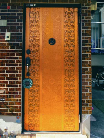 An ornate bright orange door with dark circular hardware stands within a brown brick-patterned facade.