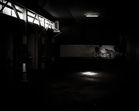 A bicycle stands against the far wall of a dark covered space with one sunlit patch in black and white.