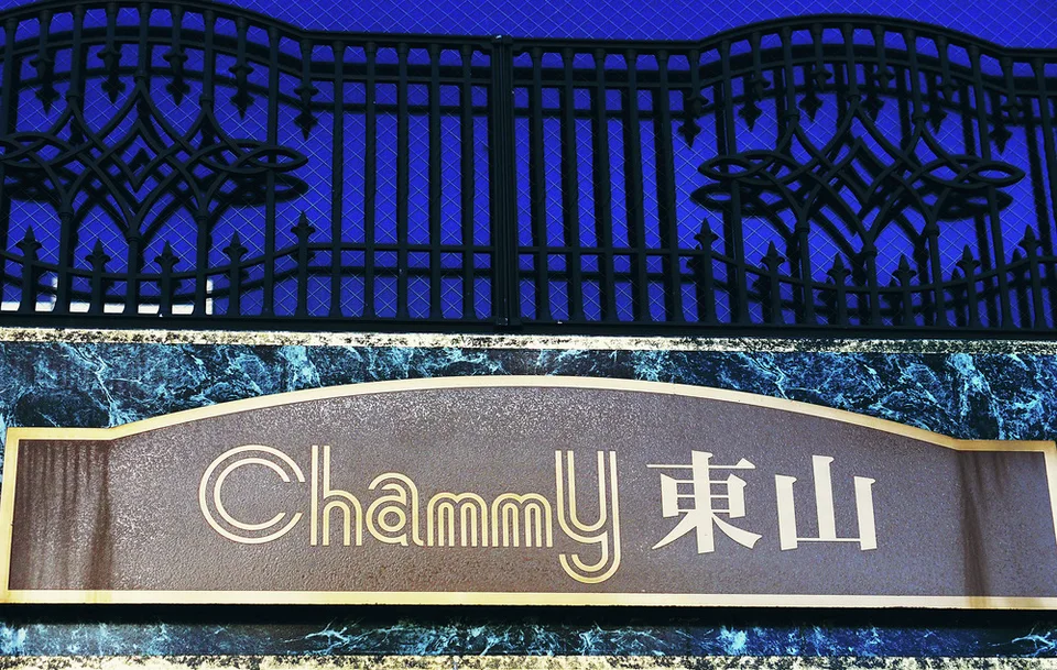 A dark gold-bordered name sign sits below an ornate black railing against a blue facade.