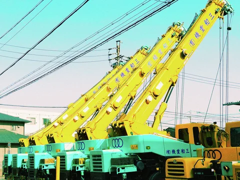 Several yellow crane booms angle above turquoise vehicles beneath crisscrossing overhead wires.