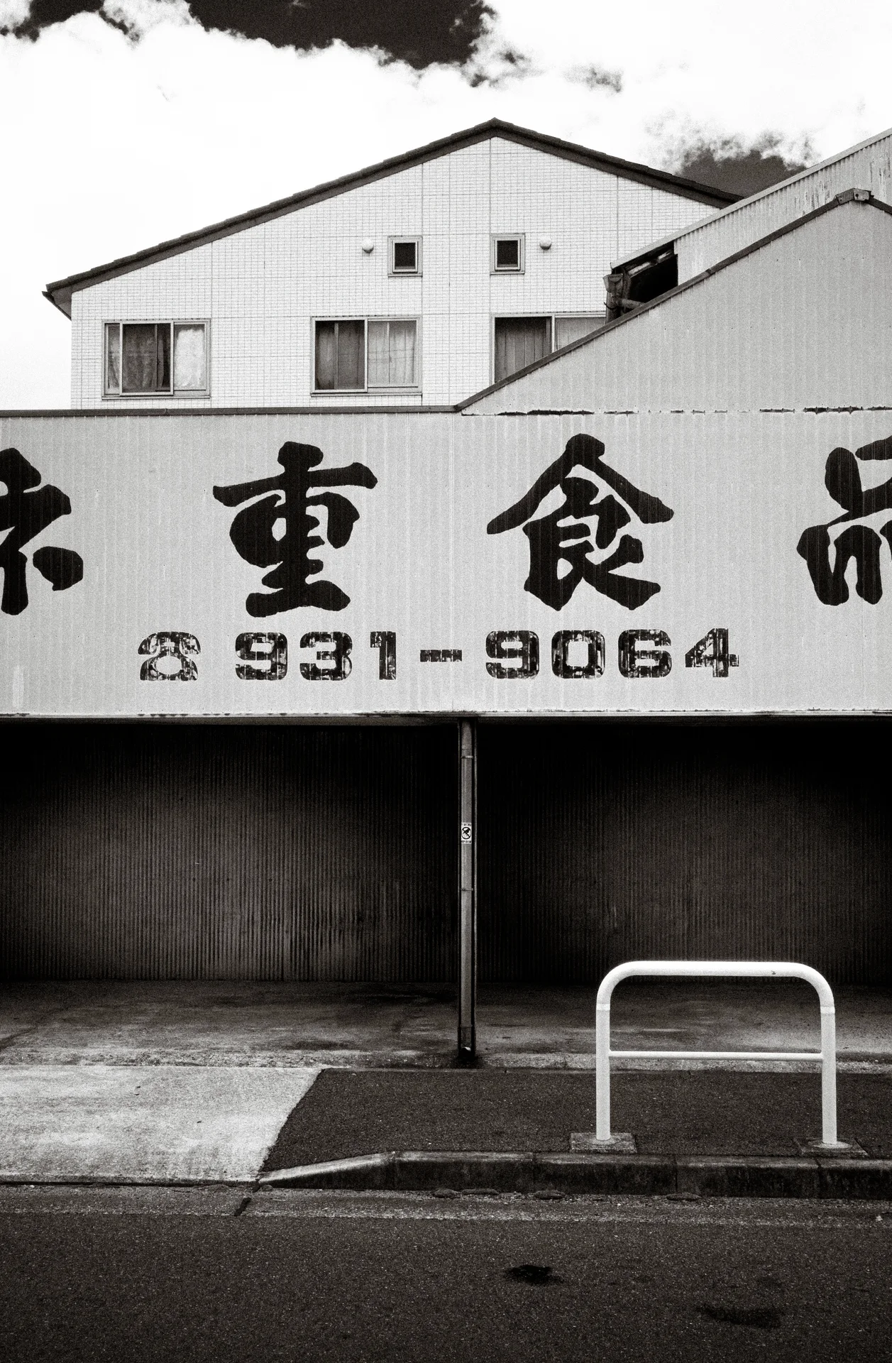 Bold Japanese lettering and telephone numbers span a low storefront sign above a roadside barrier in black and white.