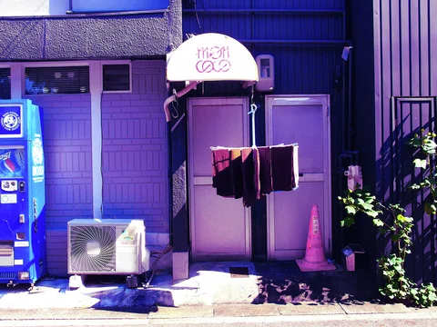 Dark cloths hang before a narrow doorway beside a blue vending machine, air conditioner, and pink cone.