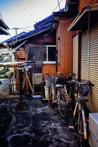 Bicycles and laundry racks line a narrow yard beside wooden houses with closed shutters and blue roof tiles.