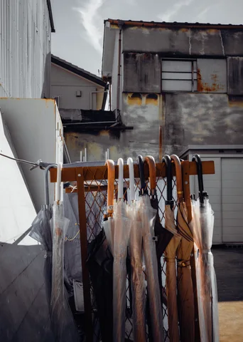 Clear umbrellas with curved handles hang from an outdoor rack before weathered industrial buildings.