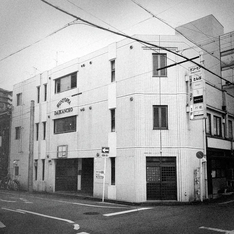 A tiled corner building has scattered narrow windows and closed street-level entrances beneath wires in black and white.