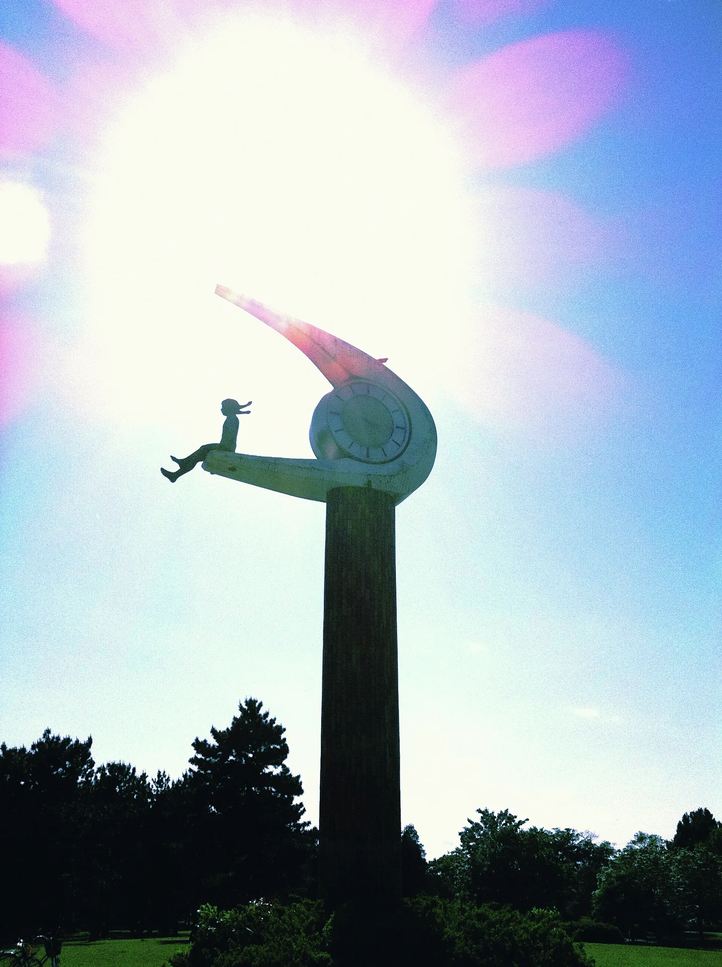 An abstract sculpture on a tall column stands silhouetted against glaring sunlight and a blue sky.