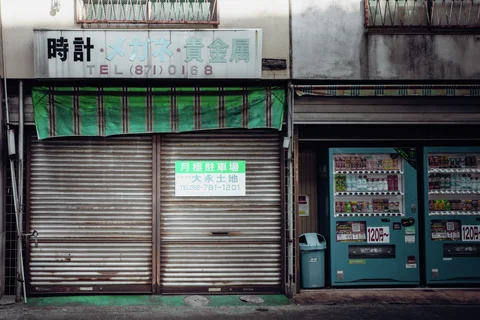 A striped green awning hangs above closed shutters beside two blue-green vending machines.