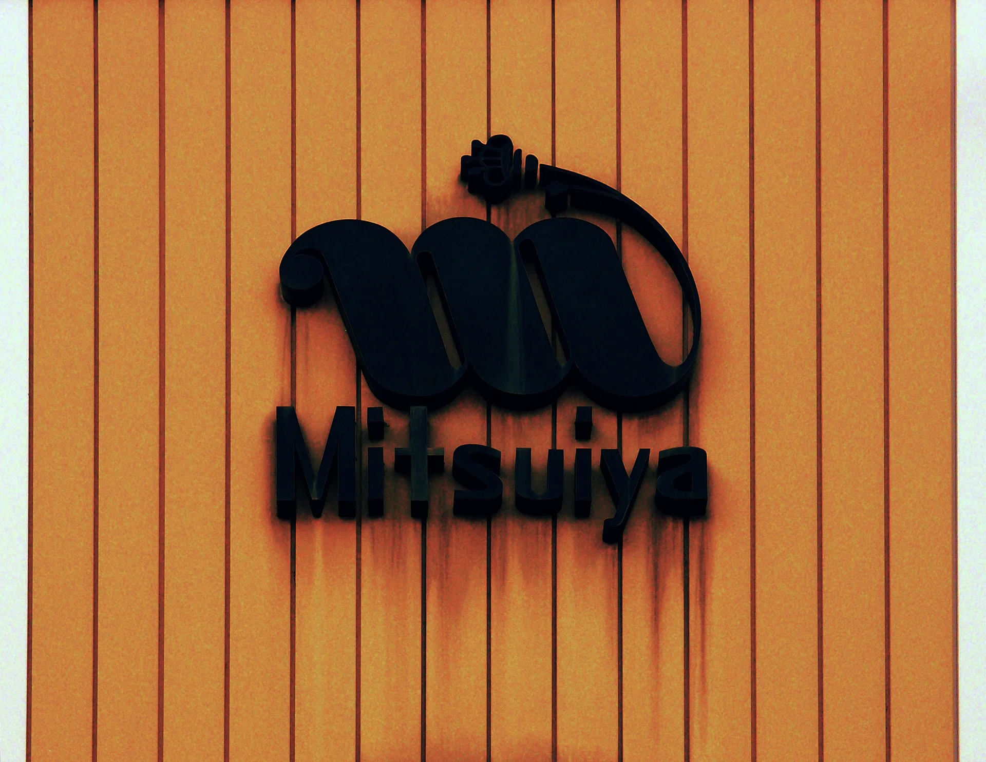 A raised black emblem and Mitsuya lettering cast shadows on orange vertical siding.