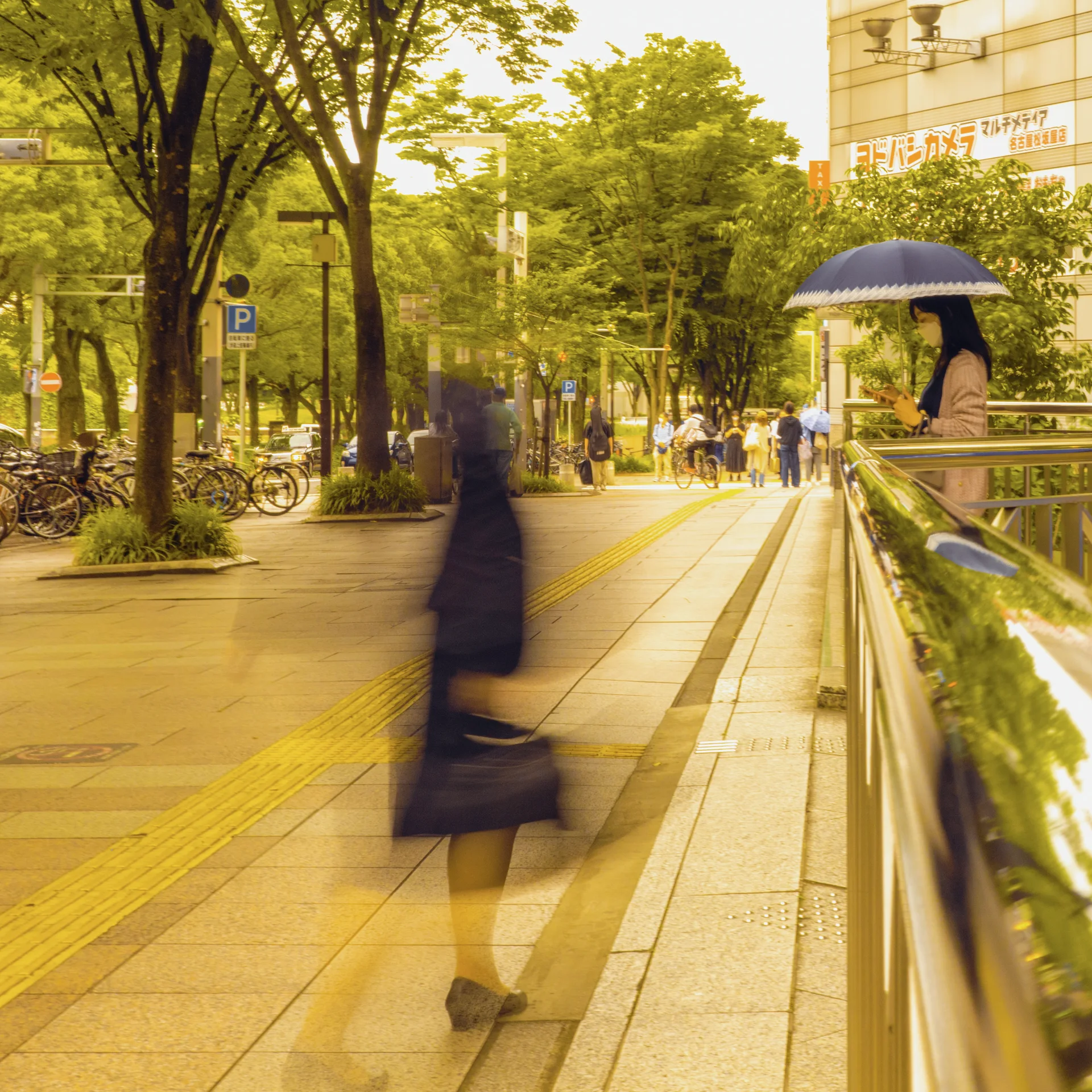 A blurred pedestrian crosses a tree-lined sidewalk beside a railing and another person holding an umbrella.