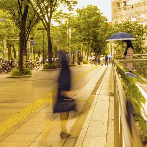 A blurred pedestrian crosses a tree-lined sidewalk beside a railing and another person holding an umbrella.