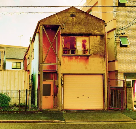 A narrow weathered yellow building has a peaked facade, boarded upper window, and closed ground-floor shutter.