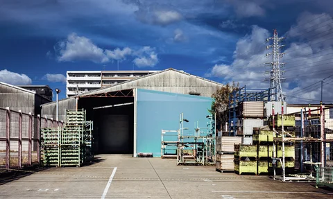 Metal storage racks line an open industrial yard before a blue warehouse beneath clouds and power lines.