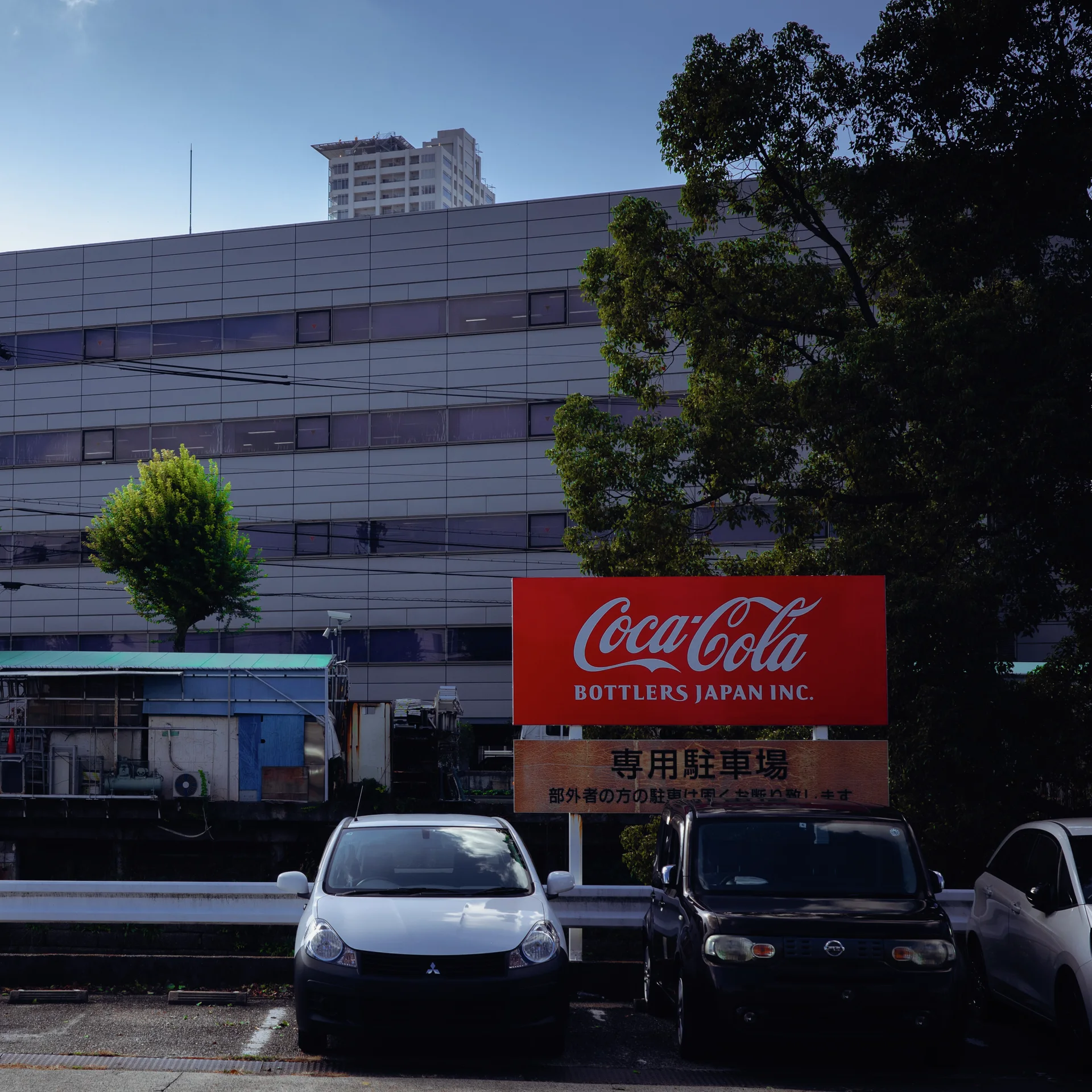 A red Coca-Cola sign stands above parked cars beside trees and a broad pale office building.