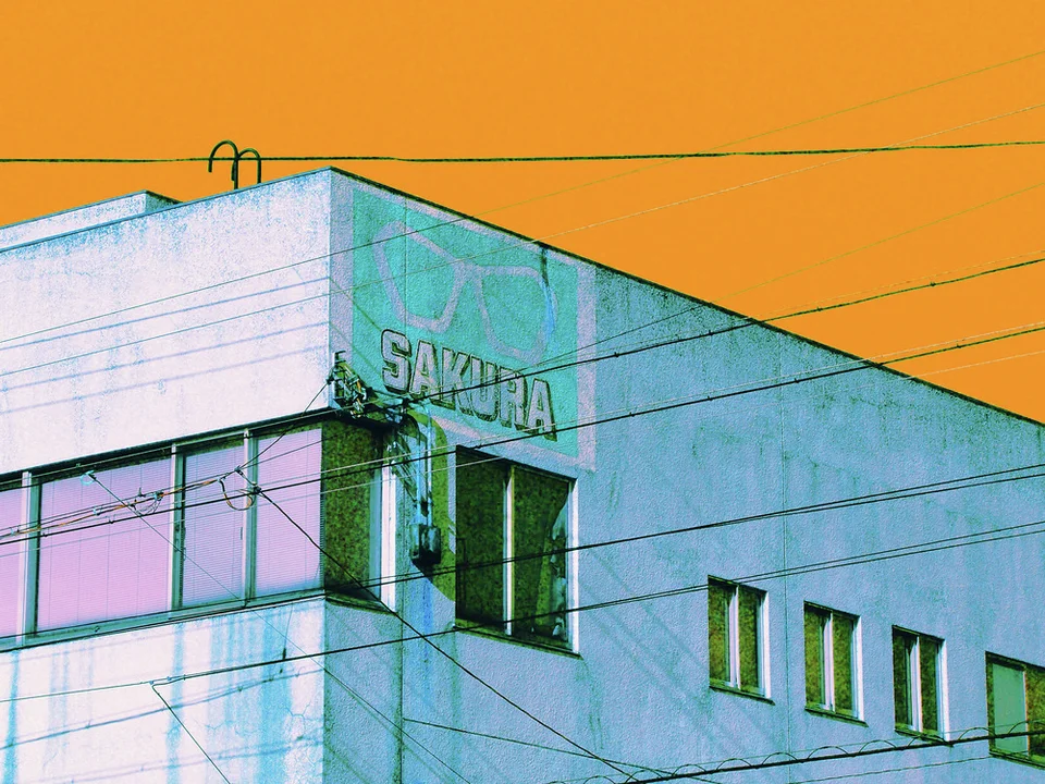 An eyeglasses symbol and SAKURA lettering mark a pale building beneath an orange sky crossed by cables.