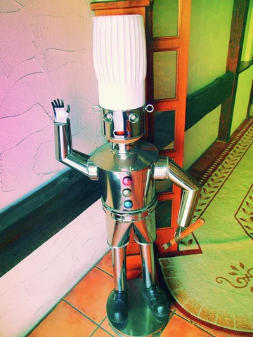 A shiny metal figure with a tall white head raises one hand beside a shop doorway.