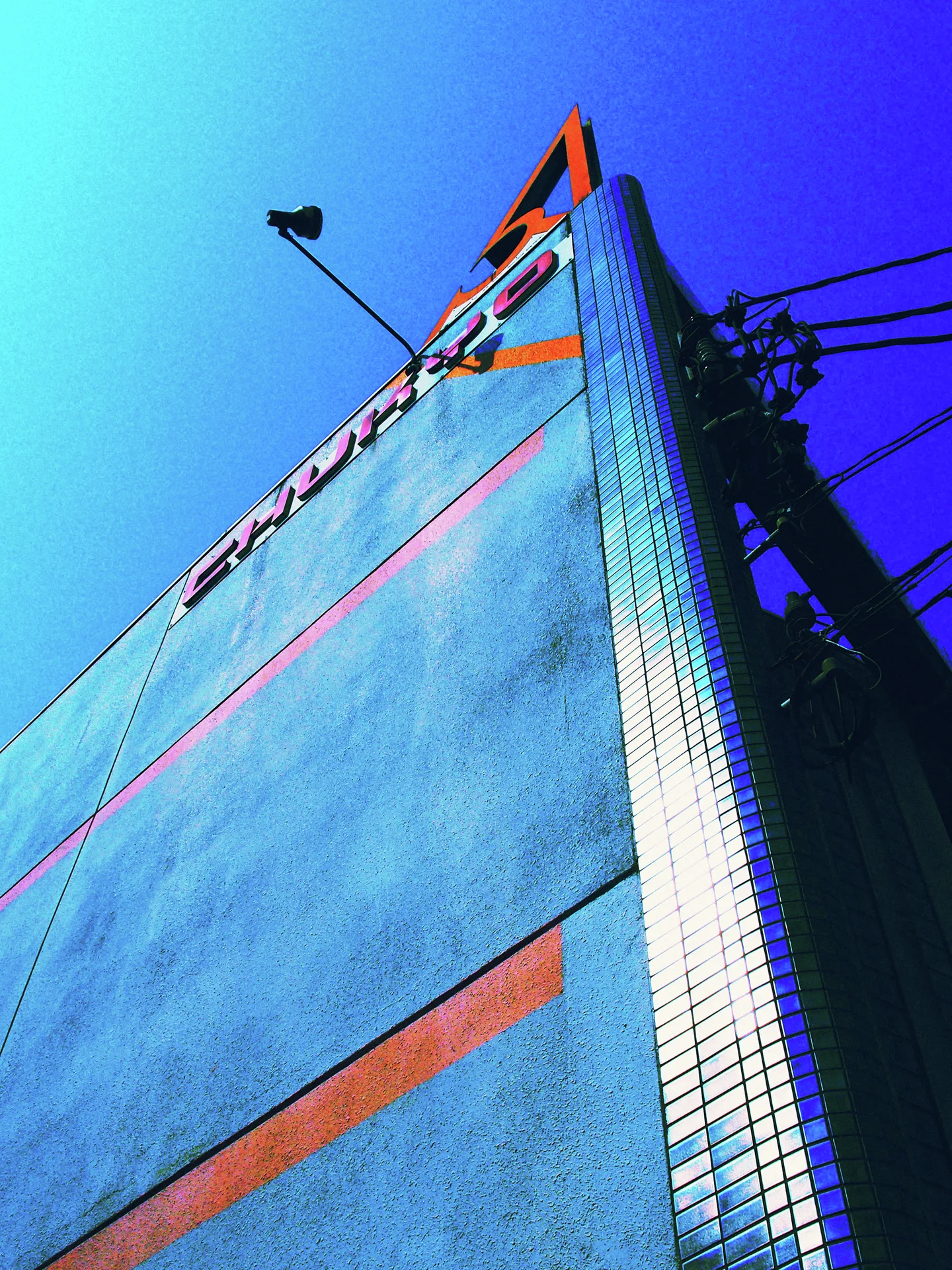 A tall blue facade with orange stripes rises toward overhead wires and bright blue sky.