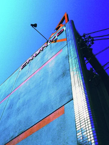 A tall blue facade with orange stripes rises toward overhead wires and bright blue sky.