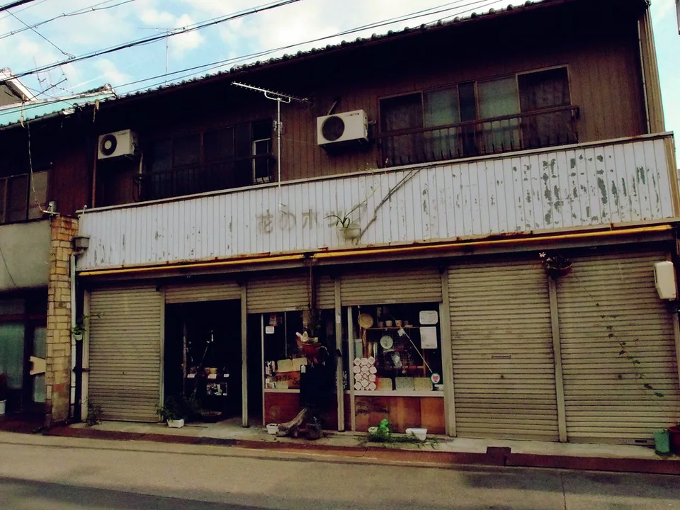 A long weathered storefront has closed shutters, small display windows, and upper air conditioners.