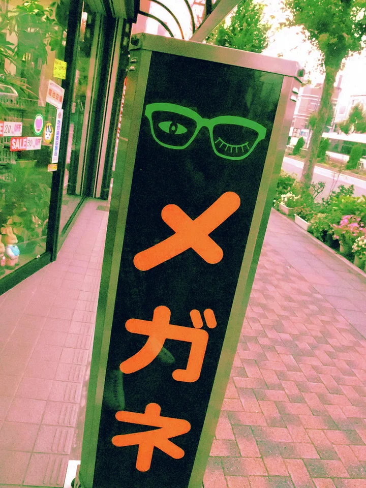 Orange Japanese lettering and a green glasses illustration decorate a narrow black sidewalk sign.
