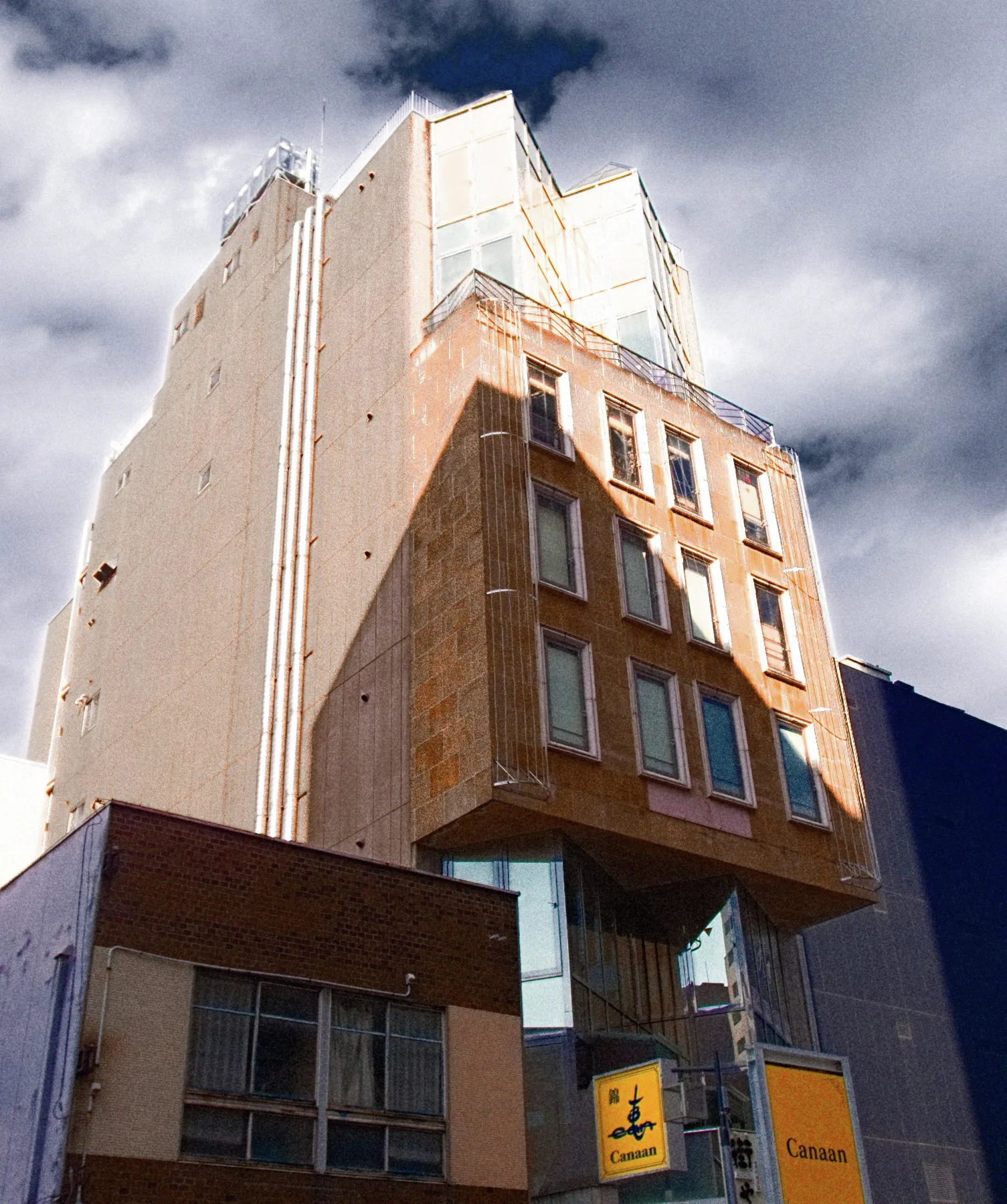 A tall pale building has a brown projecting block of windows beneath heavy gray clouds.