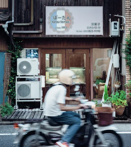 A blurred scooter rider passes a wooden storefront with stacked air conditioners, glass doors, and potted greenery.