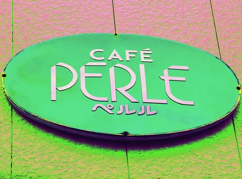 White CAFE PERLE lettering fills a green oval sign on a pink textured wall.