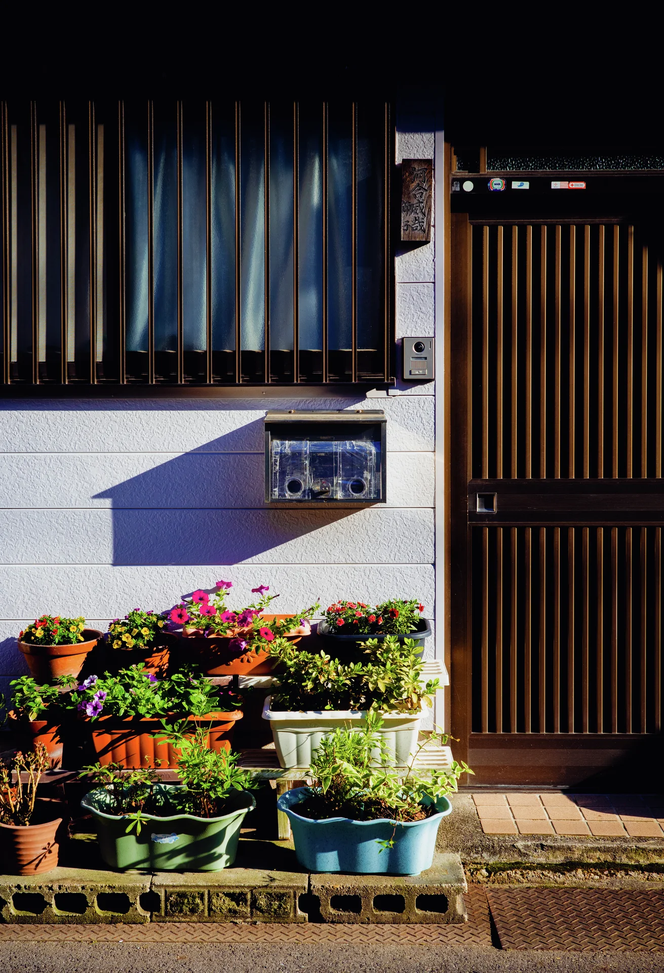 Potted flowers cluster beneath a barred window and wall-mounted mailbox beside a dark slatted entrance door.