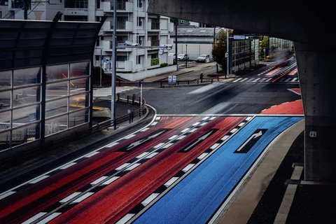 Red and blue road markings curve through an intersection beneath an elevated roadway and transparent barriers.