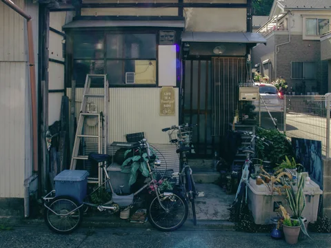 Bicycles, a leaning ladder and assorted containers crowd the small entrance of a weathered house.