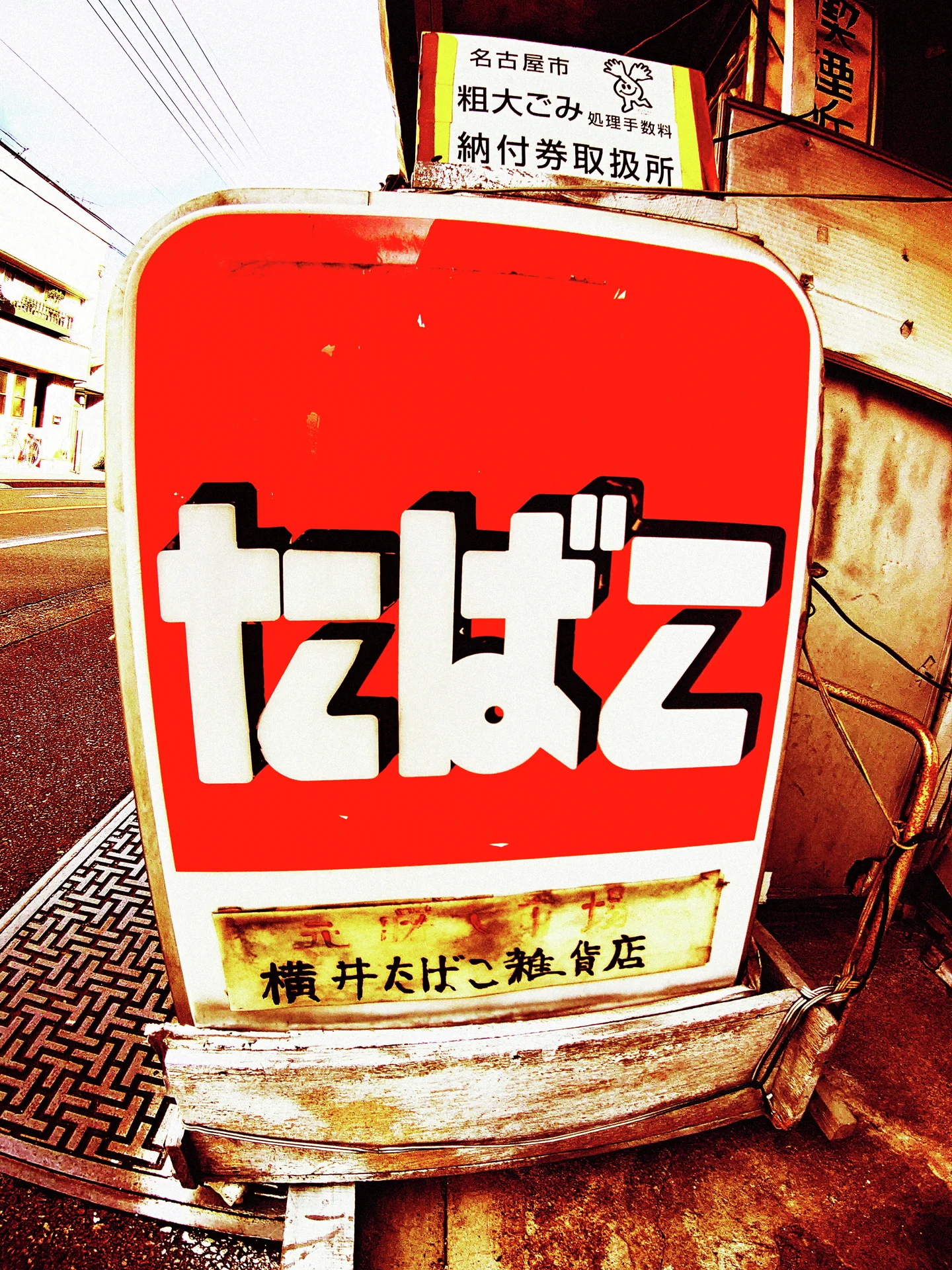 Large white Japanese lettering fills a rounded red tobacco sign above a weathered metal base.