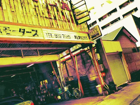 A tilted street view shows a weathered wooden storefront, stacked signs, bicycles, and a neighboring closed shutter.