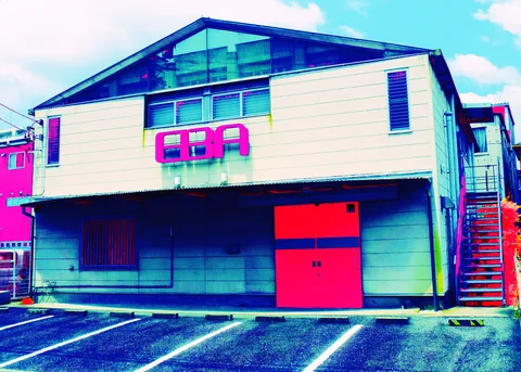 A low gabled building has a glazed roof triangle, raised pink letters, and bright red double doors.