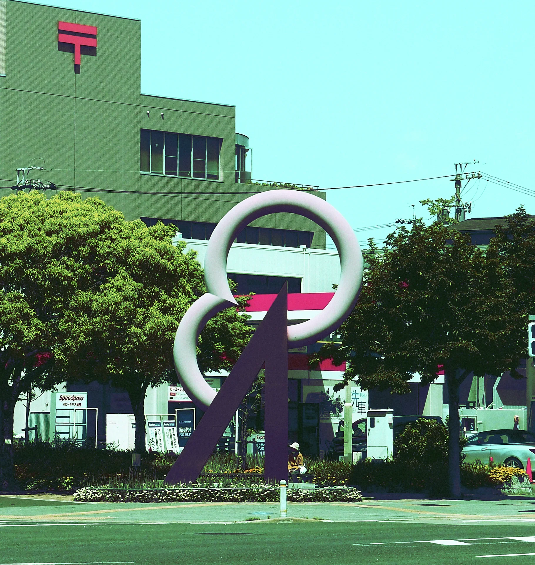 Two large pale rings balance on a dark angular sculpture among trees outside a green building.
